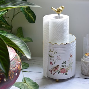 May include: A white cylindrical paper towel holder with a scalloped top edge and a gold bird finial. The exterior features a floral and bird print in muted tones. The holder stands on small gold feet and contains a roll of white paper towels.