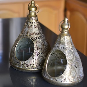 May include: Two gold-toned metal lanterns with a conical shape and intricate circular cut-out patterns. Each lantern features a circular opening at the front, a small handle at the top, and a clear glass candle holder positioned inside the base.