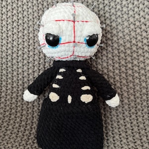 May include: A handmade crochet doll featuring a white head with red stitched grid lines and silver pins inserted throughout. The doll wears a black long-sleeved garment with white oval-shaped patches resembling a rib cage on the front torso.
