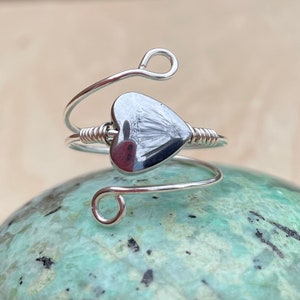 May include: A silver wire ring with a heart-shaped hematite stone. The ring is wrapped around a light green stone.