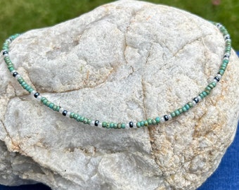 May include: A delicate necklace made with small turquoise, white, and black beads. The necklace is displayed on a light gray rock.