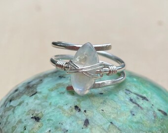 May include: A silver wire wrapped ring featuring a clear iridescent moonstone. The ring is resting on a green and black stone.