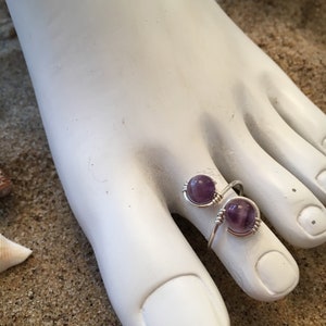 May include: A silver toe ring with two amethyst stones. The ring is on a white foot model.