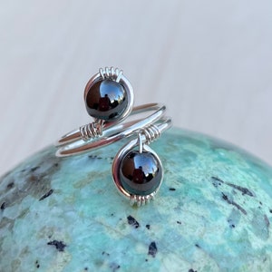 May include: A silver wire wrapped ring with two black hematite stones. The ring is on a green and black speckled stone.