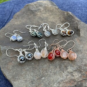 May include: A collection of gemstone earrings with wire wrapped settings. The earrings feature a variety of stones, including blue, green, grey, brown, and red. The earrings are displayed on a rock.