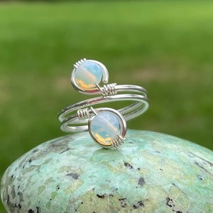 May include: A silver wire wrapped ring with two white opal stones. The ring is on a green and brown stone.