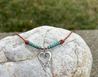 May include: A brown leather cord necklace with turquoise beads and a silver heart pendant. The heart pendant has a swirl design.