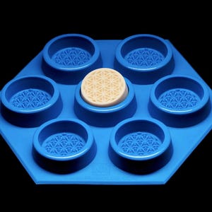 Op de afbeelding: Een blauwe zeshoekige mal met zes cirkelvormige holtes, elk met een geometrisch patroon. Een lichtgekleurde zeep met een bloem des levens-ontwerp bevindt zich in de centrale holte. De mal is gemaakt van flexibel materiaal.
