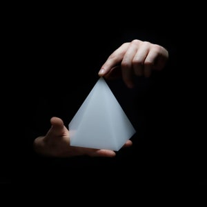 Op de afbeelding: Een wit voorwerp in de vorm van een piramide dat in de hand van een persoon wordt gehouden tegen een zwarte achtergrond.
