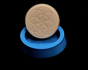 May include: A round, beige silicone mold with a raised geometric pattern, resembling a flower, on the top and bottom. The mold is sitting on a blue silicone base.