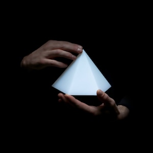 Op de afbeelding: Een witte piramide die in twee handen wordt vastgehouden tegen een zwarte achtergrond.