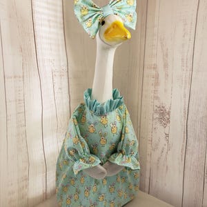 May include: A ceramic goose figurine wearing a light blue dress and bow with a yellow bunny print. The goose has a yellow beak and is wearing a ruffled collar. The dress has long sleeves and gray felt hands.