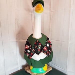 May include: A white goose figurine wearing a green cap and a green and black Christmas-themed outfit. The outfit features a pattern of nutcrackers, snowflakes, and gingerbread men. The goose is standing on a wooden base.