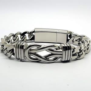 Puede incluir: Una pulsera de cadena de metal plateado con un diseño de nudo infinito central flanqueado por dos acentos cilíndricos acanalados. La pulsera incluye un cierre rectangular y muestra el texto Infinity Tributes en letra pequeña.