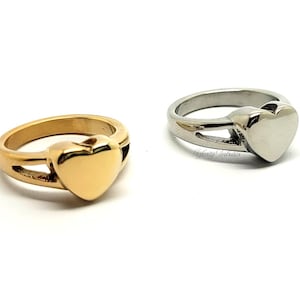 May include: Two heart-shaped rings, one gold-toned and one silver-toned, are displayed on a white background. The rings feature a heart-shaped design as the focal point, with a band that wraps around the finger. The gold ring is on the left, and the silver ring is on the right.