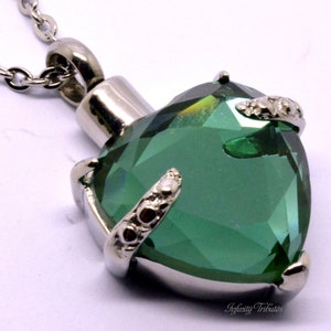 May include: A silver heart-shaped cremation pendant with a green crystal. The pendant has a detailed design with silver accents.