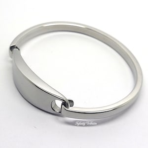 Könnte beinhalten: Ein poliertes silberfarbenes Armband mit rechteckiger Platte und kreisförmigem Verschluss. Das Armband besteht aus glattem, reflektierendem Metall und hat ein schlichtes, elegantes Design. Das Armband ist ein Schmuckstück.