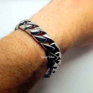 Puede incluir: Un brazalete de cadena de bordillo plateado pulido. El brazalete está hecho de eslabones grandes e interconectados. El brazalete se muestra en una muñeca. El brazalete tiene una superficie brillante y reflectante. Una pieza de joyería audaz.