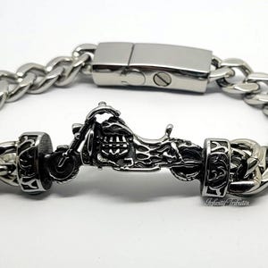Può includere: Un bracciale a maglie in metallo color argento con un ciondolo centrale a forma di motocicletta, affiancato da due elementi cilindrici decorativi con intricati motivi a volute. Il bracciale presenta una chiusura rettangolare liscia.