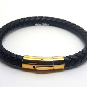 Puede incluir: Un brazalete de cuero negro con un cierre dorado. El brazalete está trenzado y tiene un diseño sencillo.