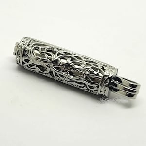 May include: A silver metal pendant with a detailed, intricate design of vines and leaves. The pendant has a cylindrical shape and a small loop at the top for attaching to a chain.