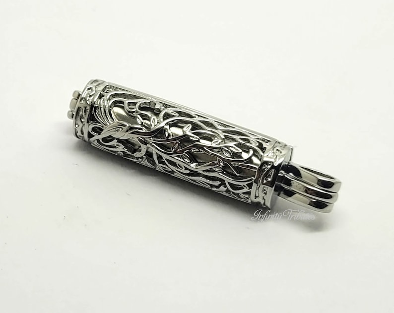 May include: A silver metal pendant with a detailed, intricate design of vines and leaves. The pendant has a cylindrical shape and a small loop at the top for attaching to a chain.