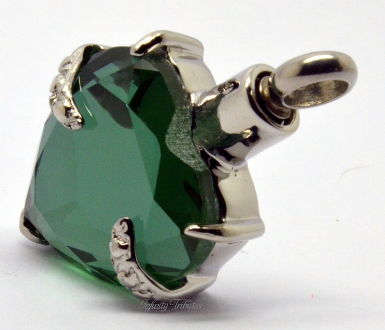 May include: A green heart-shaped cremation pendant with a silver metal frame and a small loop for attaching a chain. The pendant has a delicate floral design on the frame.
