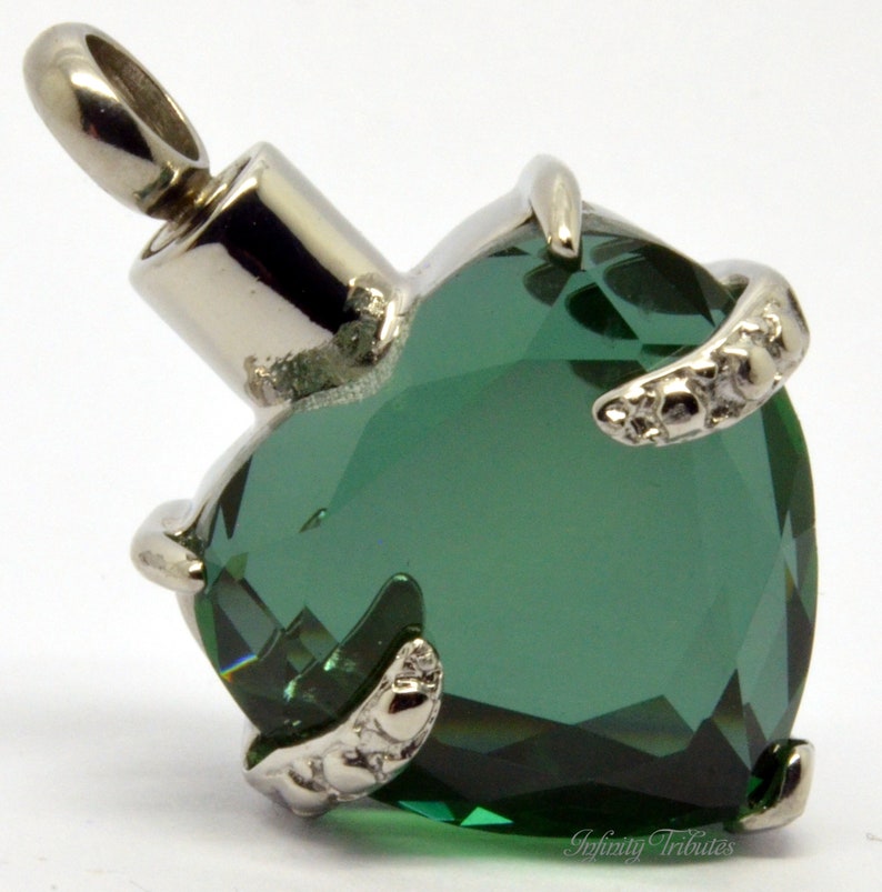 May include: A silver heart-shaped cremation pendant with a green crystal. The pendant has a small opening on the top for adding ashes. The pendant is attached to a silver chain.