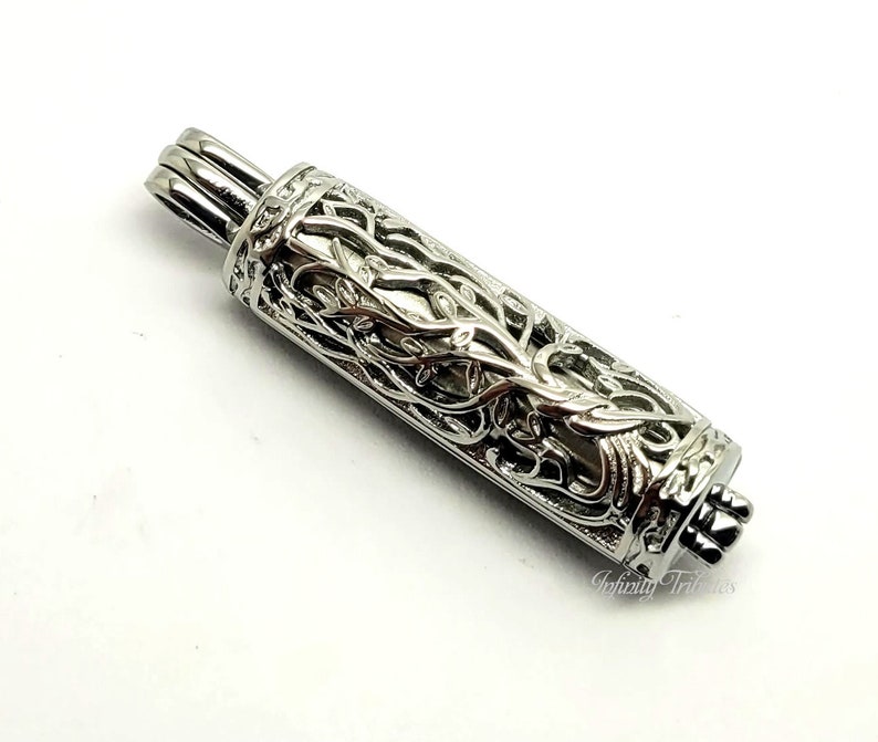 May include: Silver tone metal pendant with an intricate design of a tree with branches and leaves. The pendant is cylindrical and has a hinged lid.