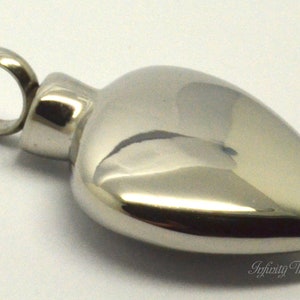 May include: A silver heart-shaped cremation pendant with a screw-on lid. The pendant has a smooth, polished finish and a loop for attaching a chain.