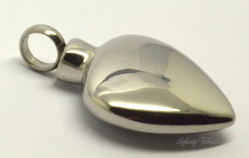 May include: A silver heart-shaped cremation pendant with a screw-on lid. The pendant has a smooth, polished finish and a loop for attaching a chain.