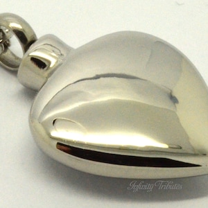 May include: A silver heart-shaped cremation pendant necklace with a chain. The pendant has a smooth, polished finish.