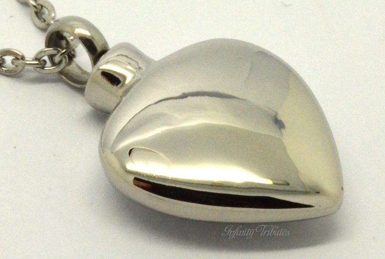 May include: A silver heart-shaped cremation pendant necklace with a chain. The pendant has a smooth, polished finish.