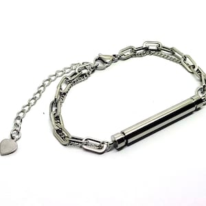 Könnte beinhalten: Ein silbernes Kettenarmband mit einem langen, zylindrischen Anhänger. Das Armband hat einen herzförmigen Anhänger am Ende der Kette.