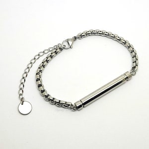 Könnte beinhalten: Ein silberfarbenes Metallarmband mit einer Box-Kette und einem zylindrischen Anhänger in der Mitte. Das Armband verfügt über einen Karabinerverschluss, eine verstellbare Verlängerungskette und einen kleinen runden Anhänger am Ende.