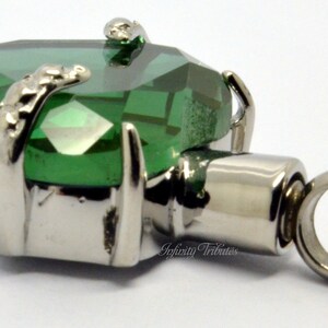 May include: A silver cremation pendant featuring a green crystal heart and a small loop for a chain. The pendant is engraved with the words "Infinity Tributes".