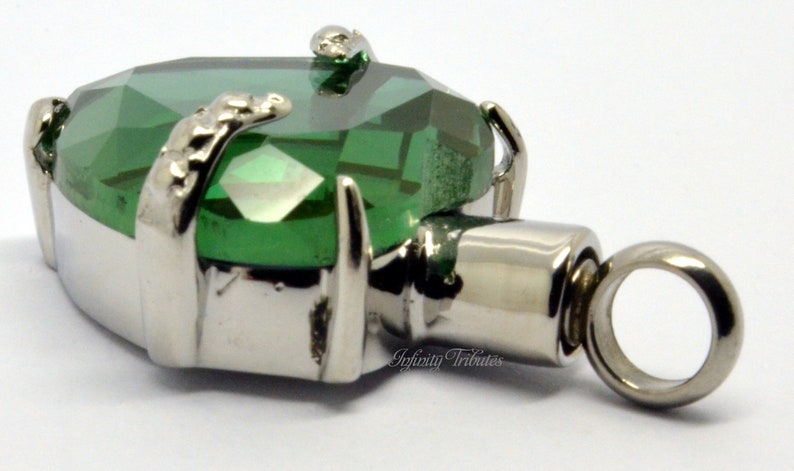 May include: A silver cremation pendant featuring a green crystal heart and a small loop for a chain. The pendant is engraved with the words "Infinity Tributes".