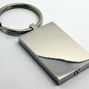 May include: A silver rectangular keychain with a smooth, reflective surface. The keychain has a ring attached to the top and a small, engraved letter "O" on the bottom.