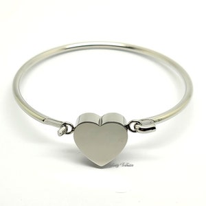 Op de afbeelding: Een zilveren armband met een hartvormige bedel. De armband is open en heeft een sluiting.
