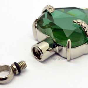 May include: A silver heart-shaped cremation pendant with a large green crystal. The pendant has a threaded screw top for opening and closing. The pendant is shown with a separate silver loop for attaching to a chain.