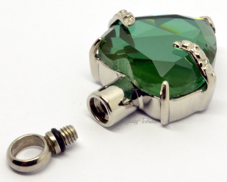 May include: A silver heart-shaped cremation pendant with a large green crystal. The pendant has a threaded screw top for opening and closing. The pendant is shown with a separate silver loop for attaching to a chain.