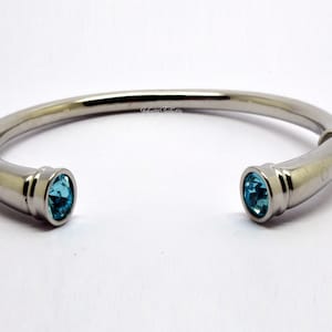 May include: A silver-toned cuff bracelet with a polished finish. The open ends feature oval-shaped, light blue gemstones. The bracelet is displayed on a white background, highlighting its sleek design and the vibrant color of the stones.