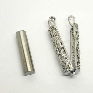 May include: A silver metal pendant with an intricate floral design. The pendant is open and reveals a hollow interior. A small silver tube is also shown.