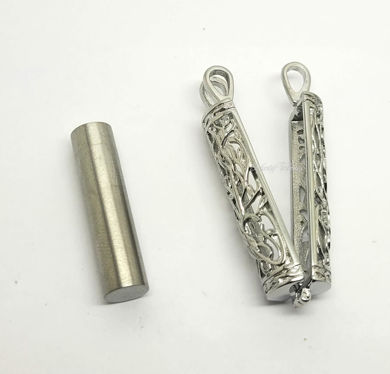 May include: A silver metal pendant with an intricate floral design. The pendant is open and reveals a hollow interior. A small silver tube is also shown.