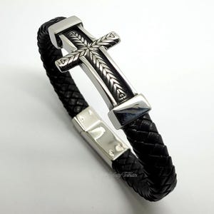 以下が含まれることがあります： 黒い革のブレスレットに、銀色の十字架のチャームが付いている。十字架は細かなデザインが施されており、銀色の留め具でブレスレットに取り付けられている。