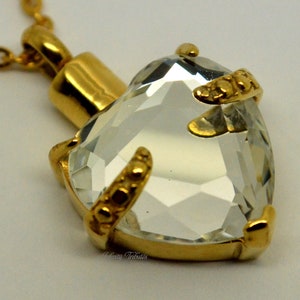 Könnte beinhalten: Goldene Halskette mit herzförmigem Anhänger und klarem Kristall. Der Anhänger hat ein detailliertes goldenes Design an den Rändern.