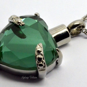 May include: A silver heart-shaped cremation pendant with a green crystal and a silver chain. The pendant has a small opening on the side for ashes.
