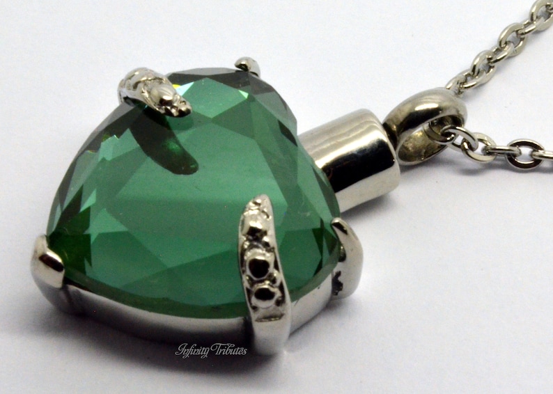 May include: A silver heart-shaped cremation pendant with a green crystal and a silver chain. The pendant has a small opening on the side for ashes.