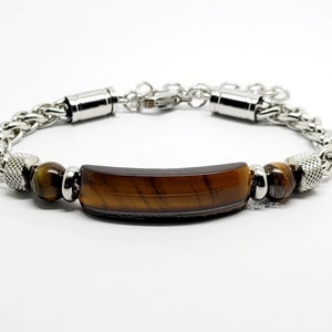 Op de afbeelding: Een roestvrijstalen armband met een schakelkettingontwerp. De armband heeft een rechthoekige tijgeroogsteen in het midden, geflankeerd door twee bruine tijgeroogkralen en zilverkleurige accenten. De armband is verstelbaar.