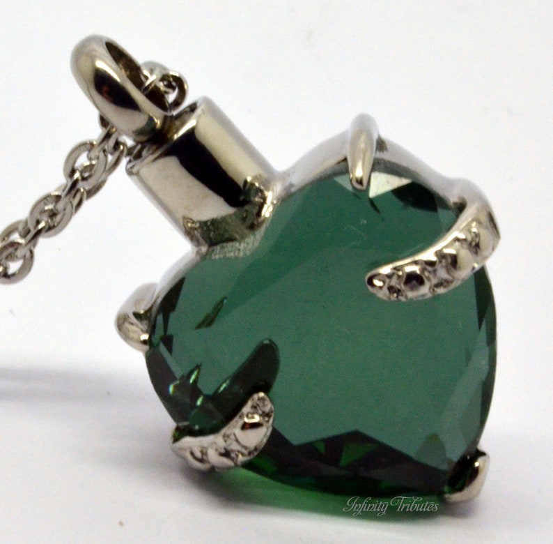 May include: A silver heart-shaped cremation pendant with a green crystal. The pendant is on a silver chain.